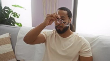 Oturma odasında sakallı bir adam, yorgun gözlerini ovuşturuyor, günlük beyaz gömlek giyiyor, kapalı bir yerde, rahat bir koltukta yorgunluk ve rahatlık gösteriyor..