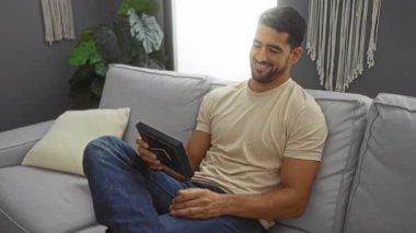 Sakallı genç adam rahat bir oturma odasında, rahat bir tişört giymiş, elinde bir fotoğraf çerçevesi tutarak, kanepede otururken gülümsüyor, bitkiler ve yumuşak dekor elementleriyle çevrili..