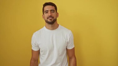 Sakallı genç bir adam, canlı sarı bir arka plana karşı el kol hareketi yapıyor. Reddediyor ve kendine referans oluyor. Ayrı bir ortamda, beyaz bir tişört giyiyor..