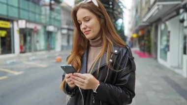 Şehirli caddede akıllı telefon kullanan genç ve çekici bir kadın. Deri ceketli, kendine güvenen bir gülümseme takınıyor..