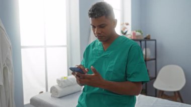 Yeşil önlüklü genç adam rehabilitasyon odasında akıllı telefon kullanıyor. Pencereden parlak bir ışık geliyor.