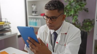 Klinikteki İspanyol erkek doktor tıbbi amaçlar için tablet kullanıyor, profesyonel bir ofis ortamında beyaz önlük ve steteskop giyiyor..