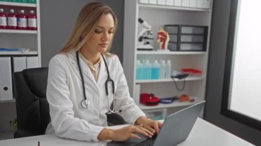Bir klinikte dizüstü bilgisayarla çalışan kadın doktor; işyeri, profesyonel bir sağlık ortamını gösteren tıbbi ekipman, şişe ve dosyalarla dolu..