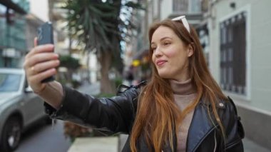Şehir merkezindeki sokak ortamında akıllı telefondan konuşan bir kadın. Şehir ortamında deri ceketli, kızıl saçlı bir kadın..