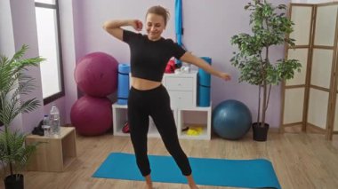 Spor salonundaki kadın ekipman ve bitkilerle neşeli bir şekilde dans ediyor, kapalı alandaki egzersiz sırasında mutluluk ve enerjiyi ifade ediyor..