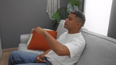 Genç adam oturma odasında bir yastıkla dinleniyor, beyaz gömlek ve kot pantolon giyiyor, kolunda bir dövme var, modern bir ev dekorunda çekilmiş..