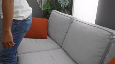 Genç adam, arka planda yeşil bitkiler ve turuncu minderlerle modern bir oturma odasında gri bir koltukta dinleniyor.