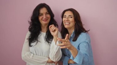 İki gülümseyen kadın pembe bir arka planda birbirlerine eşlik etmekten hoşlanırlar, mutluluğu ve sıcaklığı ifade ederler, sıradan jestleri ve neşeli ifadeleriyle..