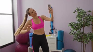 Spor salonunda gülümseyen bir kadın spor kıyafetlerini giyip spor salonunda spor topları ve bitkilerle çevrili spor kıyafetleriyle gülümsüyor..