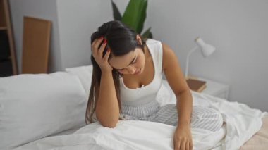 Kadın rahat bir yatak odasında, beyaz çarşaflar içinde rahat bir şekilde oturuyor ve iç mekanda, modern bir lamba ve kitaplarla birlikte düşünceli görünüyor..