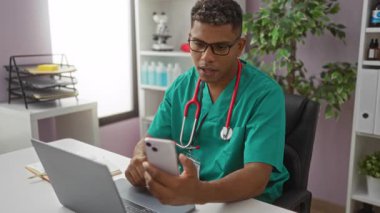 Klinikteki genç adam ameliyat önlüğü ve steteskop takıyor, akıllı telefonla konuşurken dizüstü bilgisayarla çalışıyor..
