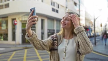 Kentsel caddede selfie çeken kadın örgü hırka giyiyor canlı şehir yaşamını canlı bir gülümsemeyle yakalıyor akıllı telefon, sarı saç ve bahar güneşinde sıradan bir tarz kullanıyor.