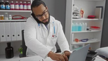 Tıbbi malzeme ve dosyalarla dolu bir klinikte, telefon ve dizüstü bilgisayar arasında steteskop kullanan beyaz önlüklü genç bir adam..