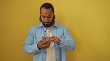 Sakallı genç adam gülümsüyor ve parlak sarı bir arkaplana karşı akıllı bir telefon tutarken başparmağını kaldırıyor, pozitifliği ve dijital bağlantıyı ifade ediyor..