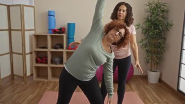 Orta yaşlı İspanyol kadınlar, spor salonunda birlikte yoga yapıyorlar. Bir kadın, egzersiz sırasında olgun arkadaşını eğitiyor ve cesaretlendiriyor..