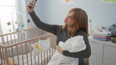 Bebek odasında selfie çeken, bebek tutan, gülümseyen, oyuncaklarla çevrili, modern annelik gösteren, ebeveynlik ve teknoloji dengesini gösteren bir kadın..