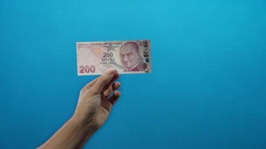 Mavi izole bir zemin üzerinde Türk lira banknotu tutan, canlı bir şekilde para ile etkileşen bir adam tasvir.