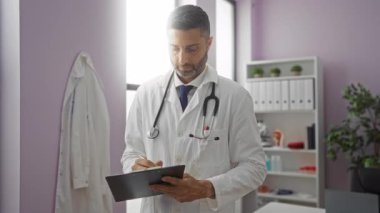 Beyaz önlüklü doktor hastane odasındaki panoya not alıyor. Arka planda tıbbi ekipmanlarla birlikte. Profesyonellik ve sağlık ortamını öneriyor..