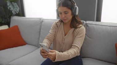 Kulaklık takan bir kadın rahat bir oturma odasında koltukta otururken akıllı bir telefonla selfie çekiyor. Evde rahatlama ve modern teknolojiyi ön plana çıkarıyor..