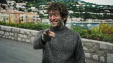 Dışarıdaki bulanık şehir manzaralı kamerayı gösteren gri kazaklı gülen adam kentsel bir ortamda spontanelik ve bağlantı olduğunu gösteriyor.