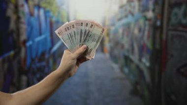 Bangladeshi Taka 'yı elinde tutan adam duvar yazısıyla, açık havada, kentsel finans temaları ve kültürel bağlamları vurguluyor.