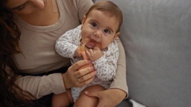 Anne evde bebeği kucağında tutarken sevgi ve bağlılık dolu bir aile anını gösteriyor. Bir kadın ve bebeğini sıcak, samimi ve içten bir ortamda ön plana çıkarıyor..