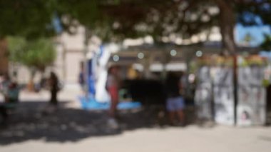 Güneşli Mallorca 'da çözünmüş bir açık hava pazarında yürüyen bulanık insanlar ve arka planda gölgeler bırakan ağaçlar.