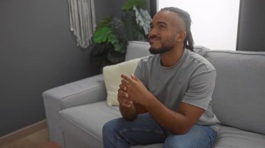 Oturma odasında örgülü genç bir adam gürültü yüzünden kulaklarını kapatıyor, bir kanepede oturuyor, bitkiler ve gri tonlarla modern bir apartman dekorunda hayal kırıklığına uğramış görünüyor..