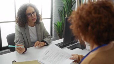 İki İspanyol kadın bir iş ortamında çalışıyor. Masasında bilgisayarla modern bir ofiste belgeleri tartışıyorlar..