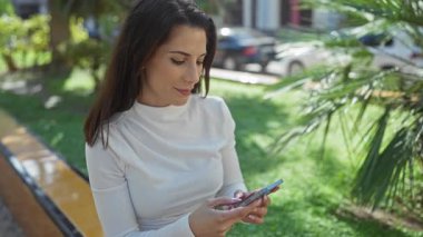 Şehir parkında akıllı telefon kullanan bir kadın güneşli bir günde arka planda arabalarla ve yeşilliklerle günlük yaşam tarzını ve bağlantısını sergiliyor..