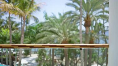 Bulanık tropikal tatil köyü sahnesi, palmiye ağaçları ve odak noktasında cam parmaklıklar bulunan, güneşli bir günde bulanık ve bokeh etkisi yaratan odaksız bir arka plan.