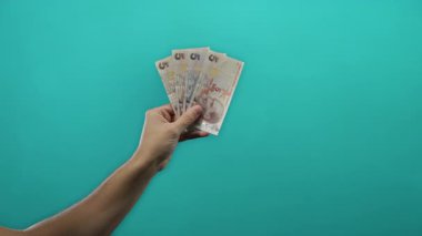 İzole edilmiş mavi duvar arkaplanına karşı elinde Türk lirası banknotları tutan adam, para, finans ve ekonomiyi basit ve net bir bağlamda sembolize ediyor.