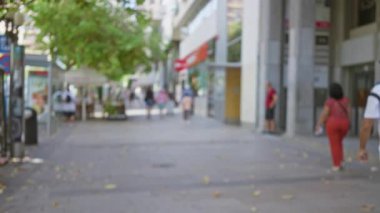 Güneşli bir günde ağaçlarla kaplı bir sokakta dükkanları ve kafeleri olan insanların bulanık görüntüsü.