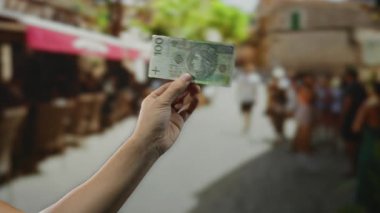 Güneşli bir günde açık hava manzarası sunan, insanların ve kafelerin bulanık geçmişine sahip, hareketli şehir caddesinde Polonyalı bir banknot tutan adam..