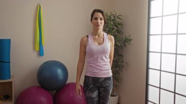 Spor salonundaki kadın spor kıyafetleri giyiyor ve arka planda spor aletleri ve yoga minderleriyle davetkar pozlar veriyor..