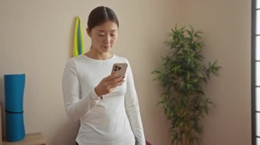 Spor salonunda spor aletleri ve yoga minderleriyle akıllı telefon kullanan bir kadın barışçıl bir ortamda modern teknoloji ve fitness karışımı bir şey yaratıyor..