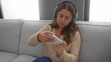 Akıllı telefonuyla oynayan bir kadın rahat oturma odası ortamında kulaklıklarla rahat ve odaklanmış bir atmosferi sergiliyor..
