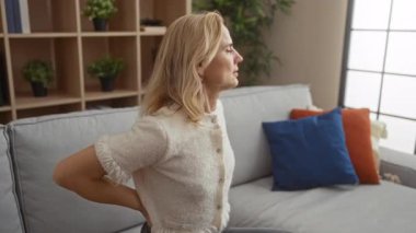 Modern bir oturma odasında, dekoratif yastıklar ve raflarla kanepede otururken sırt ağrısı çeken bir kadın, evdeki rahatsızlığını dile getiriyor..