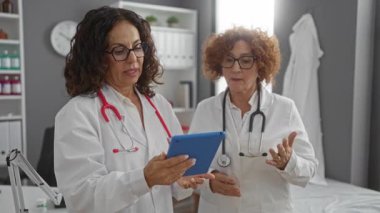 İki kadın doktor, orta yaşlı, bir hastane kliniğinde tablet kullanmayı tartışıyorlar, profesyonel ve işbirlikçi bir sağlık ortamı sergiliyorlar..