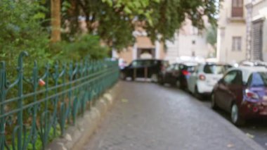 Roma 'da park halindeki arabalarla ve demir çitlerle kaplı, güneşli bir günde etrafı tarihi binalar ve yemyeşil ağaçlarla çevrili bir kaldırım taşı caddesinin bulanık görüntüsü.