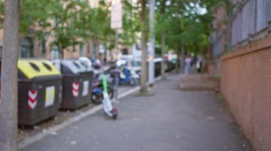 Bulanık şehir caddesi, sessiz bir Bokeh öğleden sonrasında yemyeşil ağaçların altındaki kaldırımda duran mobiletler ve çöp konteynerleriyle..