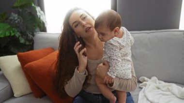 Oturma odasında bebeğini kucağında tutarken telefonla konuşan bir kadın, ev ve çevreye odaklı sevgi dolu bir aile sahnesini canlandırıyor..