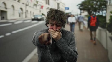 Kalabalık bir şehir sokağında neşeli bir şekilde kamerayı işaret ederek gülen genç bir İspanyol adam çevrende canlı bir şehir ortamında yürürken.