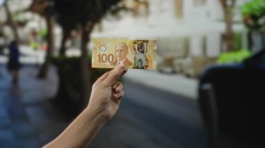 Kanadalı 100 dolarlık banknotu açık bir şehir caddesinde elinde tutuyor. Şehir yaşamını ve finansal temalarını bulanık bir şehir manzarasına karşı açıkça sergiliyor..
