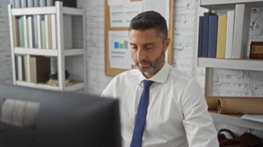 Ofisteki genç adam bilgisayar başında çalışırken boynu ağrıyan, kapalı alanda kitap rafları ve çizelgeleri olan bir ortamda rahatsızlığını ve stresi iletiyor..