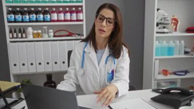 Esmer saçlı kadın doktor yorgun görünüyor ve kapalı bir klinikte tıbbi malzemeler ve ekipmanlarla çevrili bir hastane odasında masasında otururken gözlüklerini çıkarıyor..