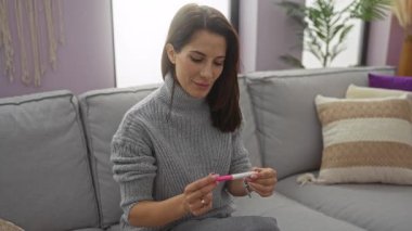 Konforlu oturma odasında hamilelik testi tutan, neşe ve şaşkınlık ifade eden, koltukta oturan, gri süveter giyen, modern iç mekanda mor aksanlı bir kadın..