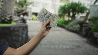 Bir şehir caddesinde Amerikan banknotlarını tutan bir adam, arka planda yeşillik bulanıklaştırılmış gündelik bir ortamda para sergiliyor, kentsel ve finansal temalar öneriyor.