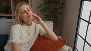 Oturma odasındaki kanepede rahat bir kadın. Yüzünde düşünceli bir ifade, pencerenin yanında ve rahat ev ortamında bitkiler var..