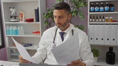 Tıbbi ekipman ve çizelgelerle çevrili bir klinikteki belgeleri inceleyen, steteskop takan ve evrak işlerine odaklanan bir doktor..